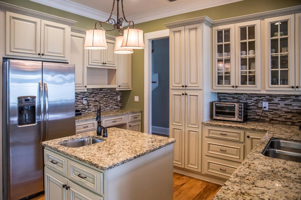A square shaped green kitchen with cream colored cabinets in a new construction home with granite countertops and lots of cabinets and storage space