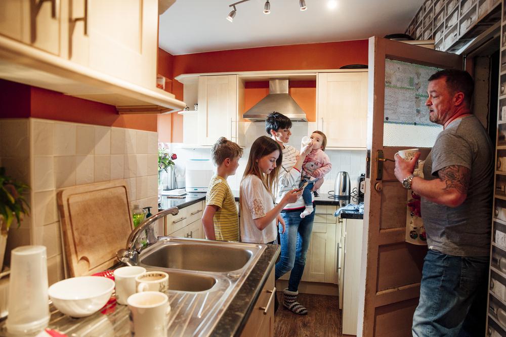 A whole family are busy in the kitchen of their home. The mother is feeding the baby while the father does the dishes. The older girl is using a smartphone and her brother is watching the baby get fed.